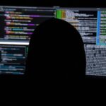 A person in silhouette against multiple computer monitors displaying lines of code