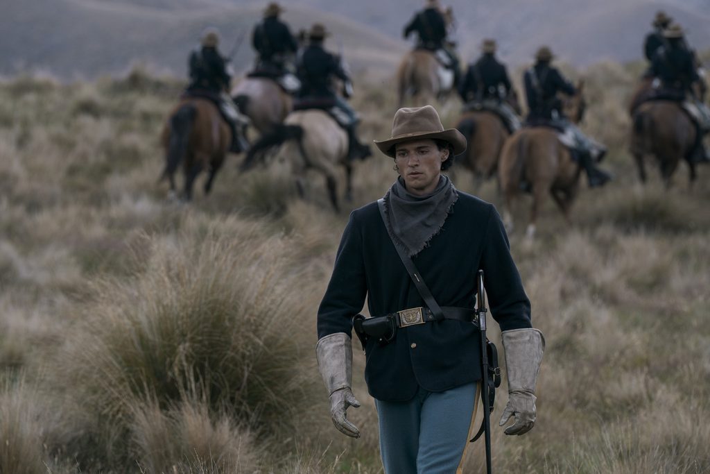 "A man in historical cavalry uniform walks through grassy plains with mountains in the background, while a group of mounted riders moves away behind him."
