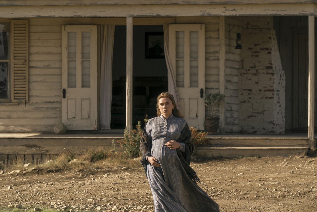"A woman in a long dress stands outside a weathered, old wooden house with open doors and shutters, surrounded by dry dirt and sparse plants, under natural daylight."