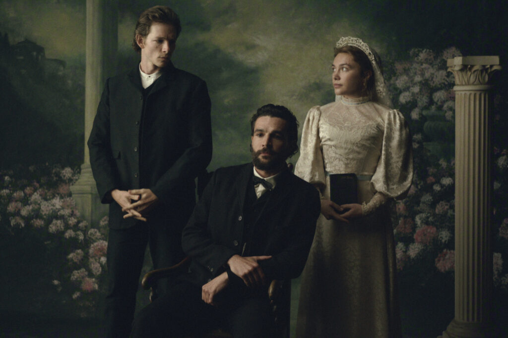 "Three people in Victorian-era clothing pose in a formal portrait setting with floral and classical column background; one man seated, one man standing, and a woman in a white dress holding a book."