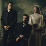 "Three people in Victorian-era clothing pose in a formal portrait setting with floral and classical column background; one man seated, one man standing, and a woman in a white dress holding a book."