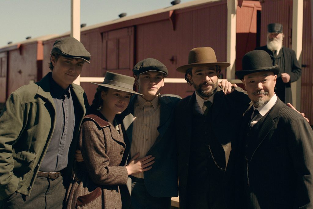 "Five people in vintage clothing pose together outdoors by a red train car, with clear skies above and a wooden platform. The setting and attire suggest an early 20th-century or historical period scene."